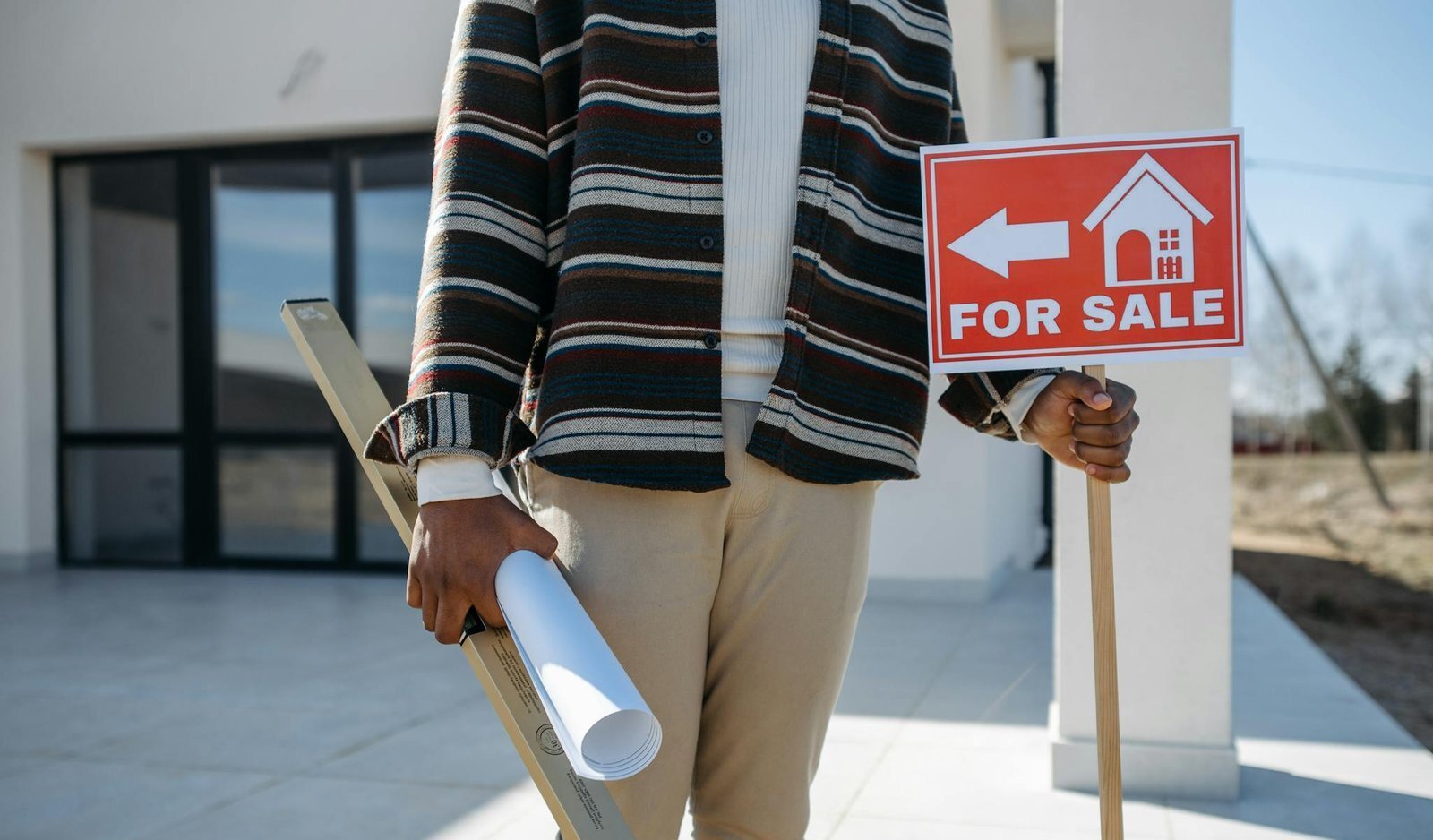 A real estate agent stands outside with a 'for sale' sign and rolled blueprint, suggesting a property for sale.